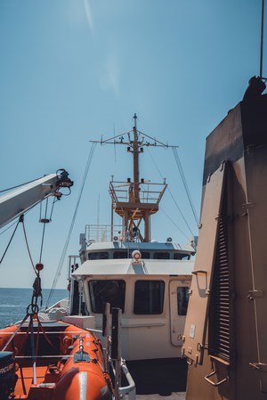 Vanaf het dek van een schip zie je de witte brug met mast en antennes, een oranje reddingssloep en een crane onder een strakblauwe hemel boven zee.