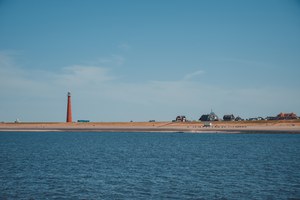 Een rode vuurtoren rijst op boven een langgerekte kustlijn met verspreide huizen, gezien vanaf het kalme water onder een strakblauwe lucht.