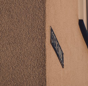 Een blauw geëmailleerd straatnaambordje op de hoek van een gebouw met ruwe pleisterwand en een gladde, warmbeige muur.