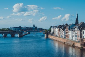 Rijtjeshuizen langs de Maas met een oude boogbrug op de achtergrond en een kerktoren die boven de skyline uitsteekt onder een blauwe lucht.