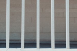 Verticale witte metalen spijlen van een hek staan voor een lichte betonnen muur van betonblokken, met een grijze stoep onderaan.
