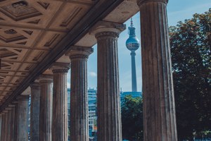 Klassieke zuilengalerij met een geornamenteerd plafond op de voorgrond, waarachter de Berlijnse Fernsehturm oprijst tegen een blauwe hemel met bomen.