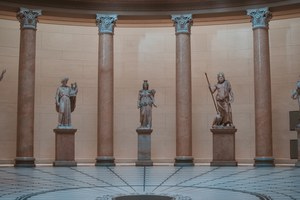 Klassieke marmeren beelden staan op sokkels tussen hoge Korinthische zuilen in een halfronde museumzaal met een geometrisch betegelde vloer.
