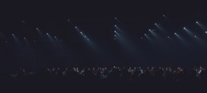 Rijen witte spotlights schijnen diagonaal door de duisternis boven een silhouet van een publiek tijdens de audiovisuele show Sora by Nonotak.