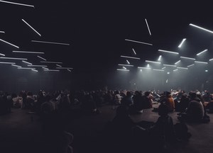 Een groot publiek zit in het donker op de vloer onder een plafond vol zwevende lichtstrepen tijdens een lichtinstallatie.