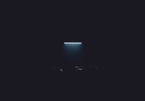 Een fel verlichte horizontale lichtbalk gloeit boven een donkere ruimte waarin de vage silhouetten van toeschouwers nauwelijks zichtbaar zijn.