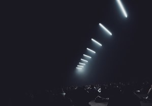 Een rij diagonaal opgehangen witte lichtstralen doorsnijdt de donkere ruimte boven de silhouetten van een publiek tijdens een audiovisuele voorstelling.