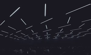 Talrijke felle lichtbuizen zweven op verschillende hoogtes onder een donker plafond boven de silhouetten van een publiek in een zaal.