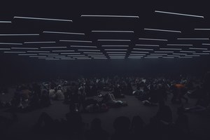 Bezoekers zitten en liggen verspreid op de vloer van een donkere ruimte onder een raster van horizontale lichtstrepen aan het plafond.