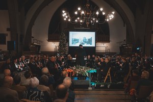 Een dirigent leidt een orkest tijdens een kerstconcert in een sfeervol verlichte kerk, met een kerstboom, projectiescherm en aandachtig publiek.