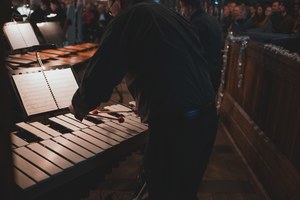 Een percussionist speelt met mallets op een marimba, met bladmuziek op de standaard en een aandachtig publiek op de achtergrond tijdens een kerstconcert.