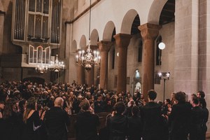 Publiek en koorleden applaudisseren in een sfeervol verlichte kerk met romaanse bogen, een groot orgel en hangende kroonluchters.