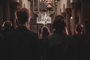 Publiek van achteren gezien in een sfeervol verlichte kerk kijkt naar een verlichte kroonluchter en orgel tijdens een concert van Kamerkoor JIP.
