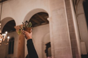 Een omhoog gestoken arm houdt een potplantje vast in een sfeervol verlichte kerkruimte met stenen bogen en een kaarsenkroon.