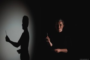 Een persoon in donkere kleding houdt een voorwerp vast in een dramatisch belicht decor, terwijl de schaduw groot op de witte wand valt.
