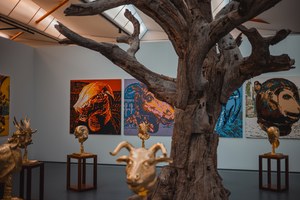 Een grote kale boom domineert een tentoonstellingsruimte met gouden dierenkoppen op sokkels en kleurrijke schilderijen van dieren aan de muren.
