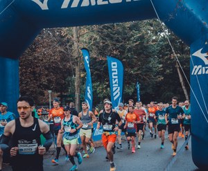 Hardlopers passeren de blauwe Mizuno-startboog tijdens de Amsterdam Marathon, met bomen en wapperende vlaggen op de achtergrond.