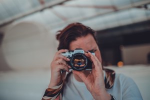 Een persoon houdt een vintage Olympus-camera voor het gezicht en richt deze op de kijker, met een lichte, wazige industriële ruimte op de achtergrond.