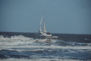 Een zeilboot met bemanning trotseert de ruwe, schuimende golven van de Noordzee onder een bewolkte grijsblauwe hemel bij Middelkerke.