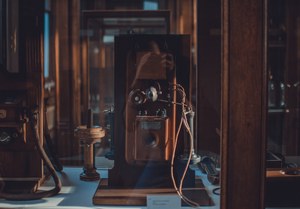 Een antieke houten wandtelefoon met koperen onderdelen en gekrulde bedrading staat in een vitrine, omringd door meer historische toestellen in warm licht.