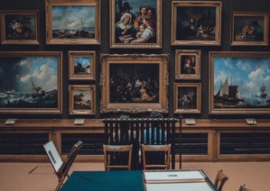 Museumzaal met donkergroene wanden vol vergulde schilderijen in klassieke lijsten, met houten stoelen en een tafel met groen kleed op de voorgrond.