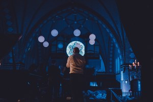 Silhouet van een man in een verduisterde kerk met verlichte bollampen, een gloeiend glas-in-loodraam en kaarsen tijdens een ligconcert.