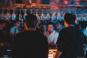 Achter een bar met rijen omgekeerde wijnglazen praten mensen in warm en blauw licht, terwijl twee mannen op de voorgrond in silhouet staan.