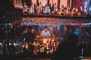 Barman kijkt op zijn telefoon achter een goedgevulde bar terwijl gasten in het warme, sfeervolle licht van een drukke cocktailbar staan.