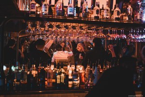Bezoekers kletsen en drinken aan een sfeervolle bar met warme verlichting, omringd door talloze flessen en boven hen hangende wijnglazen.
