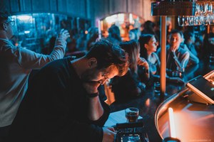 Man leunt voorovergebogen aan een bar met een glas bier, terwijl op de achtergrond mensen in blauw en oranje licht samenkomen.