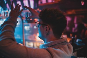 Man met bril en baard van achteren gezien, met opgeheven handen, tegen een sfeervolle achtergrond met blauwe en roze verlichting.