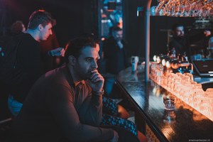 Man met baard zit peinzend aan een sfeervol verlichte bar terwijl op de achtergrond meerdere mensen in blauw-oranje licht met elkaar praten.
