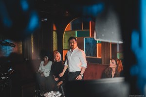 Vier mensen poseren in een sfeervolle ruimte met glas-in-loodramen, waarbij een man in wit overhemd centraal staat tussen drie zittende vrouwen.