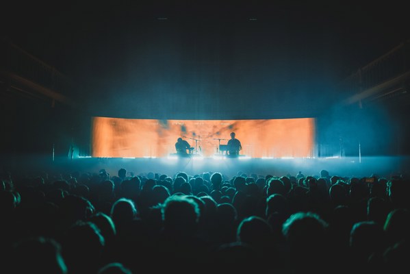 Een foto van achterin de zaal van Paradiso. We zien het publiek van achteren en de silhouetten van Weval. Ze worden van achter belicht door een LED-scherm.