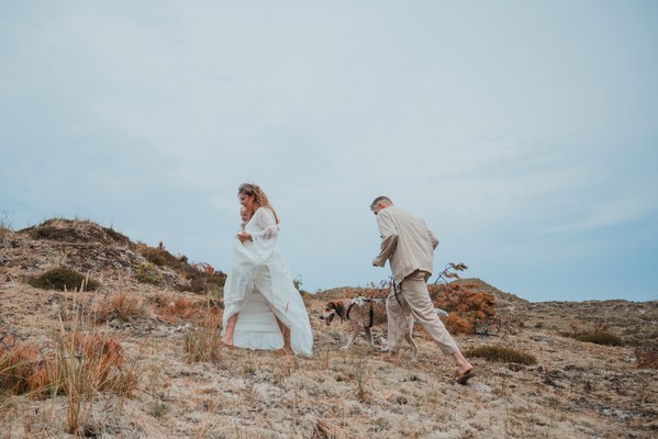 Bruid in witte jurk en bruidegom in beige pak lopen met hun hond over een droge, glooiende heuvel onder een bewolkte hemel.