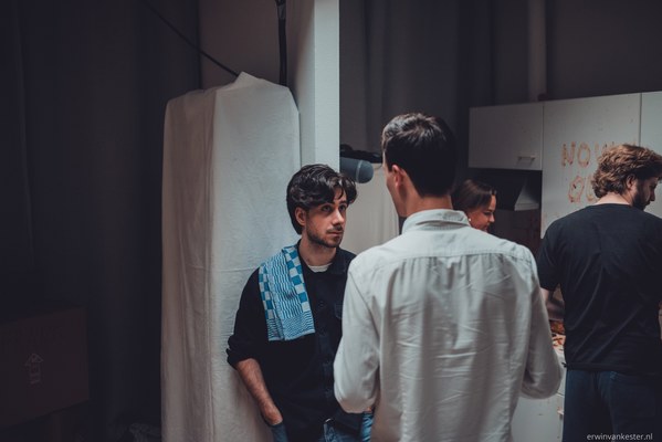 Twee jonge mannen staan in gesprek in een sfeervol verlichte studioruimte, terwijl op de achtergrond enkele andere personen bij een tafel bezig zijn.