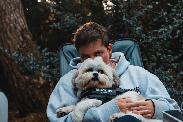 Een vrouw (Veerle) in een lichtblauwe hoodie zit in een campingstoel en houdt een hond (Kip) met Julius-K9-tuigje beschermend tegen zich aan tussen de bomen.