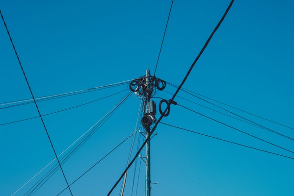 Een houten telefoonpaal met opgerolde kabels en isolatoren waaruit talloze draden uitwaaieren tegen een strakblauwe hemel.