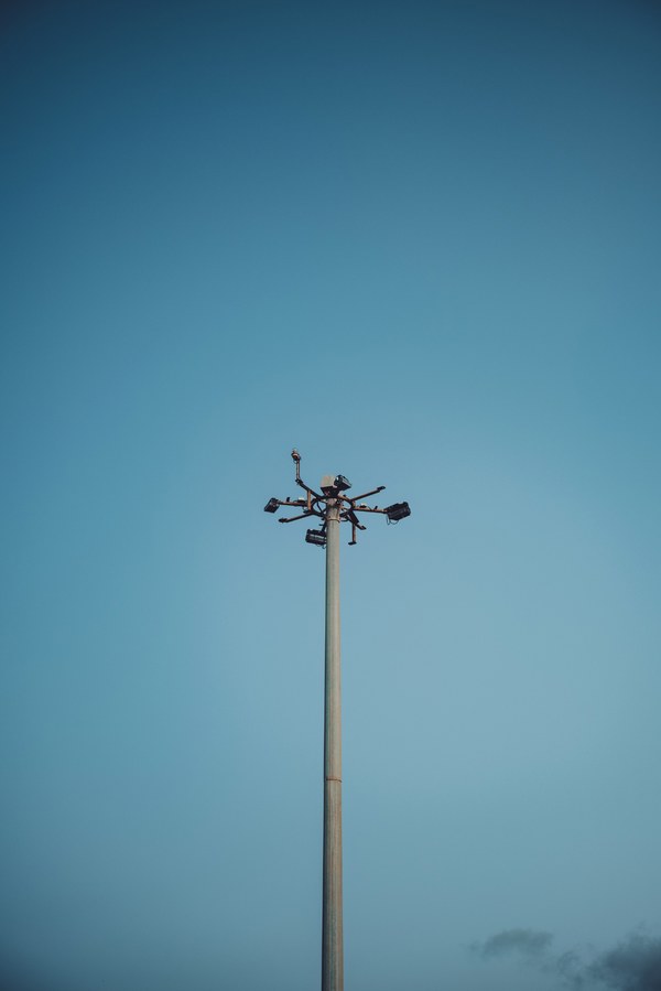 Een hoge lichtmast met meerdere schijnwerpers steekt eenzaam af tegen een diepblauwe avondhemel, met onderaan een vage wolkensluier.