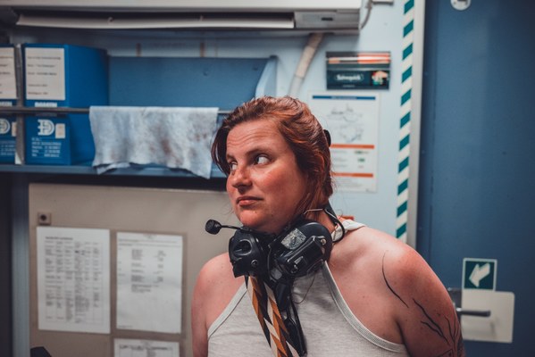 Vrouw met rood haar en een headset om haar nek kijkt opzij in een technische ruimte met kasten en documenten aan de wand.