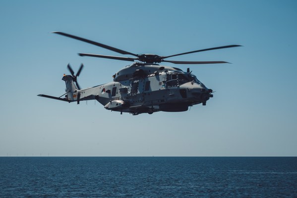 Een grijze militaire NH90-helikopter vliegt laag boven een kalme, diepblauwe zee onder een strakblauwe hemel, met windturbines vaag aan de horizon.