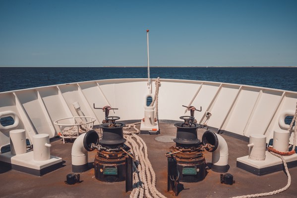 Het voordek van een schip met twee zwarte ankerlieren, opgerolde meertrossen en een witte reling, met daarachter een kalme blauwe zee onder een strakblauwe hemel.