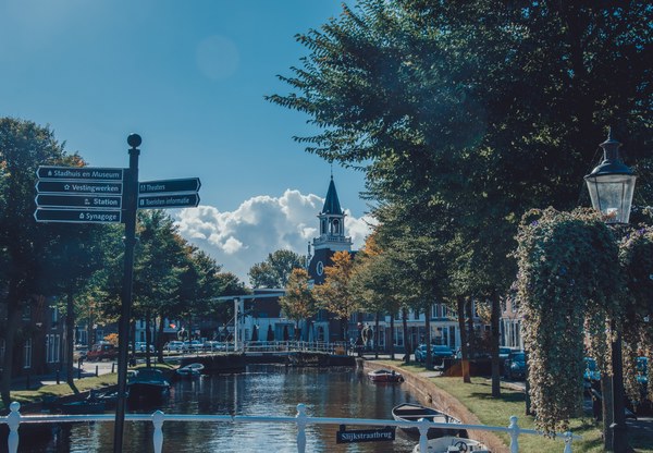 Een zonnige gracht in Weesp met een witte brug, aangemeerde bootjes, een oude kerktoren en wegwijzers naar onder meer stadhuis en station.