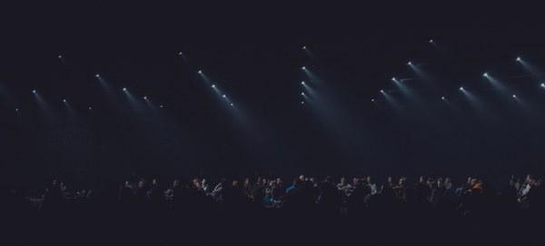Rijen witte spotlights schijnen diagonaal door de duisternis boven een silhouet van een publiek tijdens de audiovisuele show Sora by Nonotak.