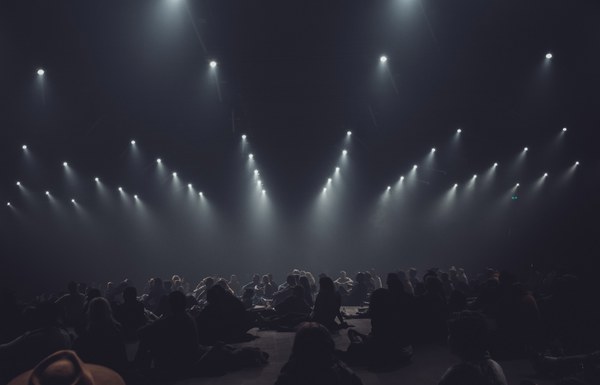 Silhouetten van een zittend publiek in een donkere zaal, verlicht door rijen felle spots die V-vormig neerstralen door de mist.
