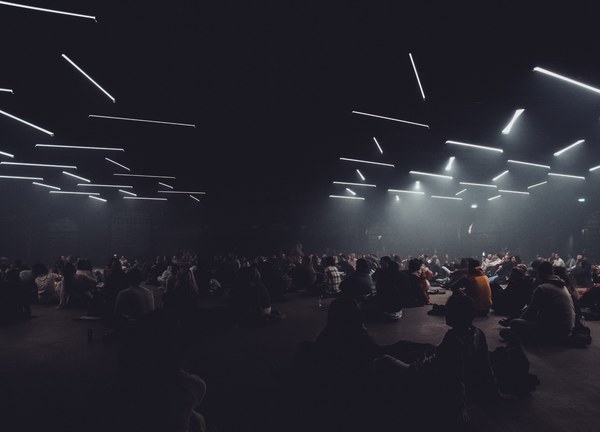 Een groot publiek zit in het donker op de vloer onder een plafond vol zwevende lichtstrepen tijdens een lichtinstallatie.