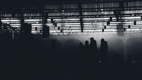 Silhouetten van mensen staan in een mistige, industriële hal met een verlicht plafond vol lichtbalken, in zwart-wit vastgelegd.