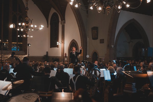 Een dirigent leidt met opgeheven armen een orkest tijdens een kerstconcert in een sfeervol verlichte kerk met kroonluchters en bogen.