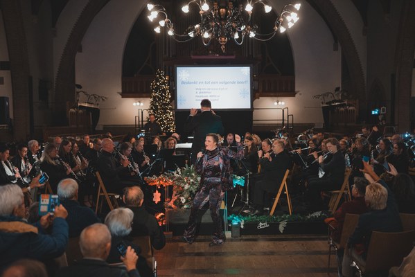 Een dirigent leidt een orkest in een sfeervol verlichte kerk tijdens een kerstconcert, terwijl het publiek toekijkt onder een grote kroonluchter.