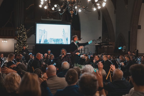 Een dirigent leidt met gespreide armen een orkest tijdens een kerstconcert in een kerk, terwijl publiek op de voorgrond meeklapt onder sfeervolle kroonluchters.